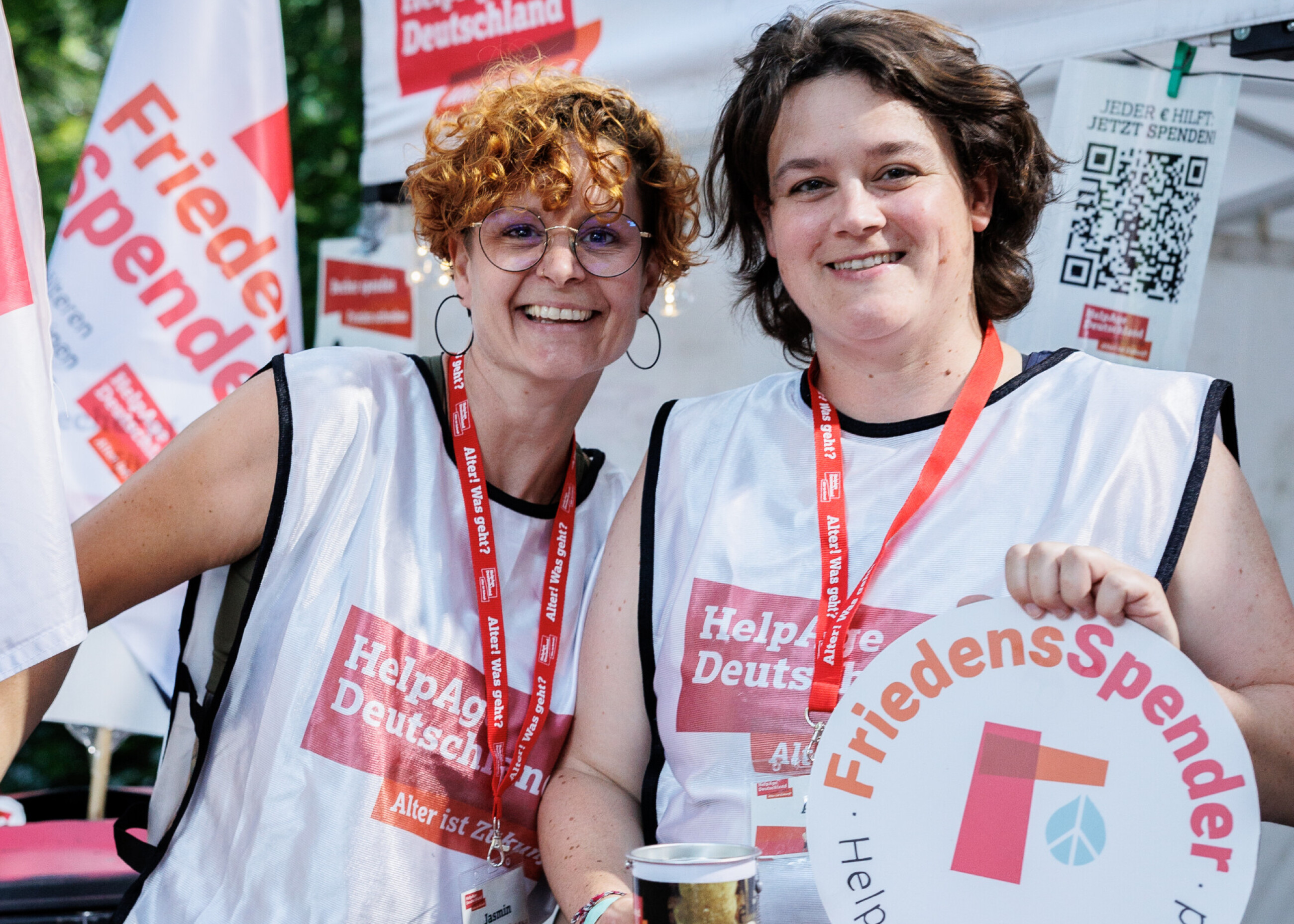 Zwei Personen gucken in die Kamera und halten das Logo vom FriedensSpender hoch