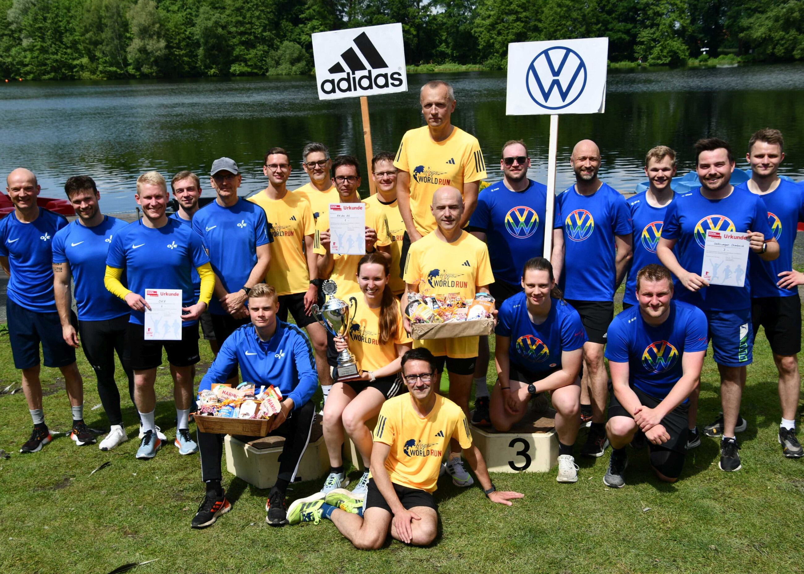 Sieger vom Firmenstaffellauf stehen von einem See und halten Schilder in die Luft